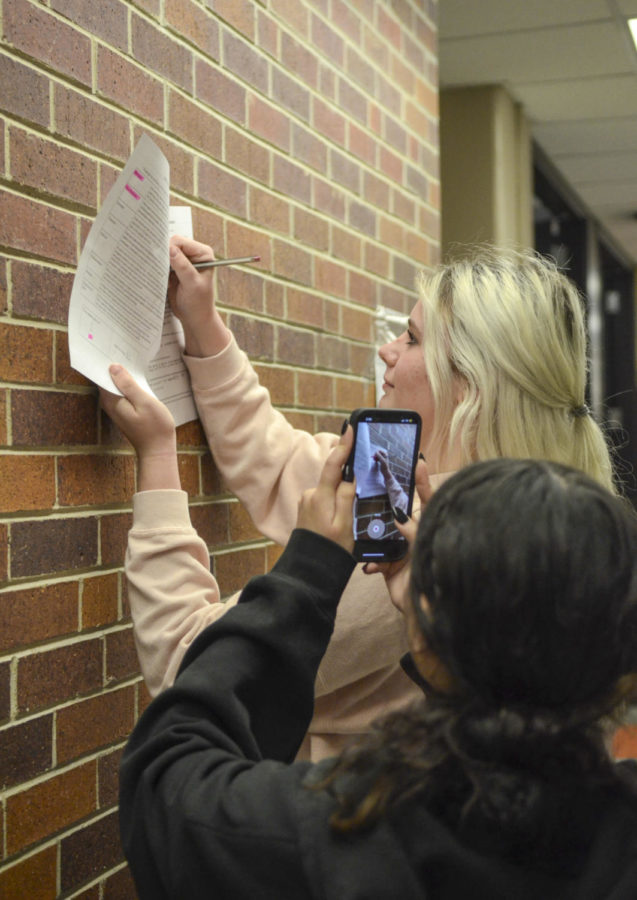 Posing, junior Savannah Kessinger and Natalia Ramirez make a TikTok for Spirit Club Jan. 19 outside of Room 101. Ramirez filmed Kessinger writing on a paper to promote turning in the Spirit Executive Board sign-ups. “We made a TikTok [account] to help promote the school as a whole on a platform that we know most of our students are on every single day.” Kessinger said.